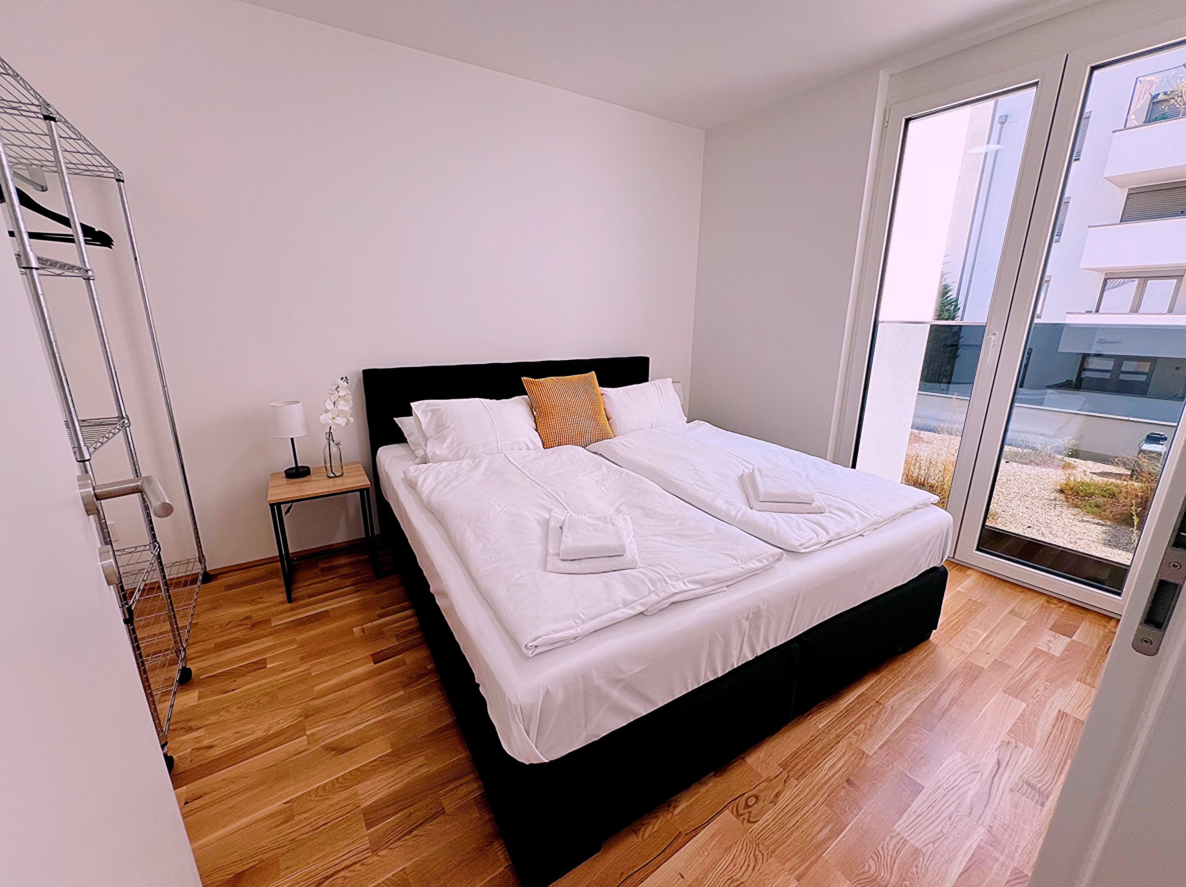 Un dormitorio moderno con una cama grande, una mesita de noche, una lámpara y vistas al exterior a través de un ventanal.
