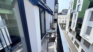 Un balcon étroit avec une petite table et des chaises, donnant sur une rue bordée d’immeubles.