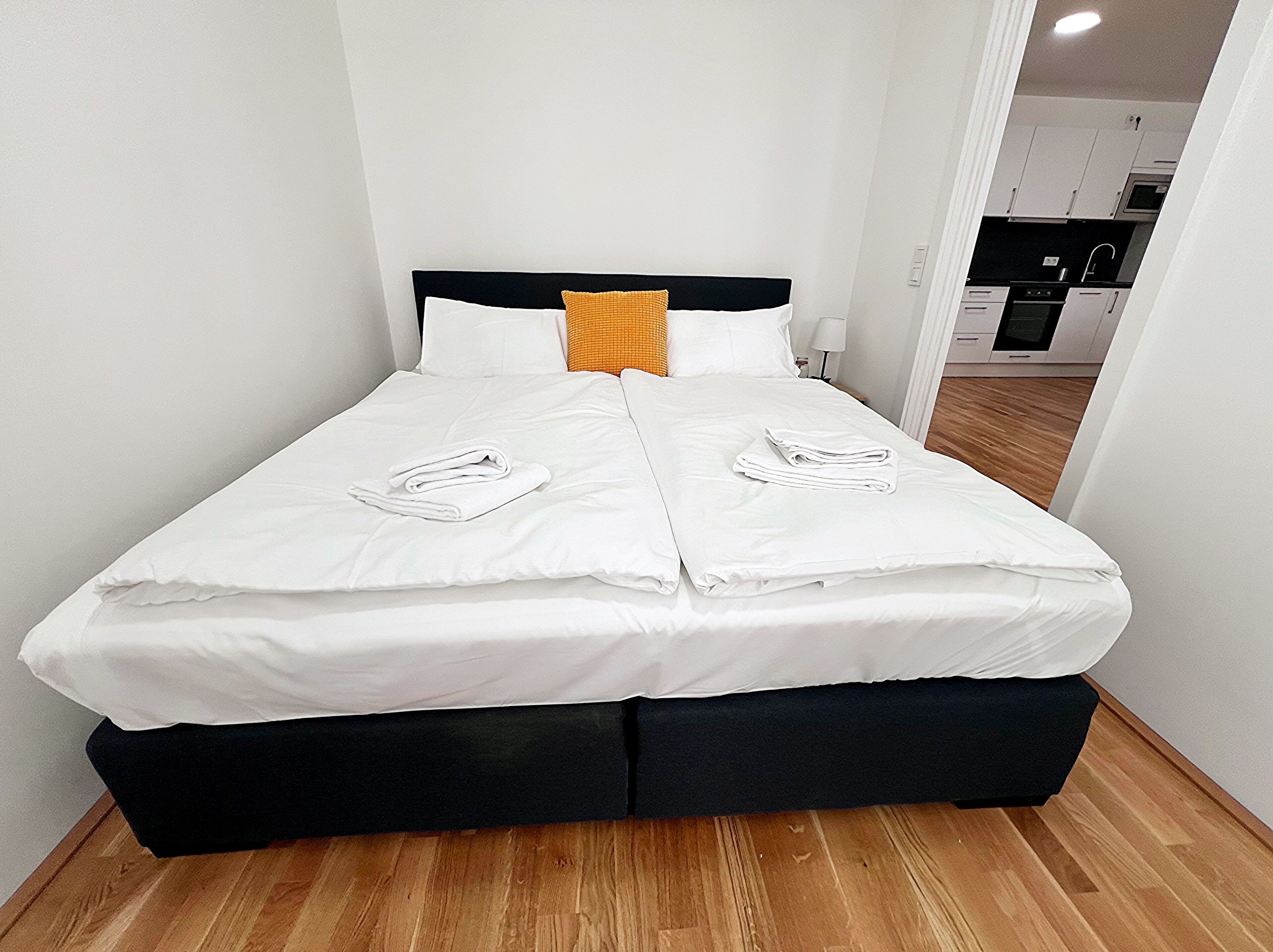 Una cama con sábanas blancas, dos toallas, un cabecero negro y una almohada amarilla.
