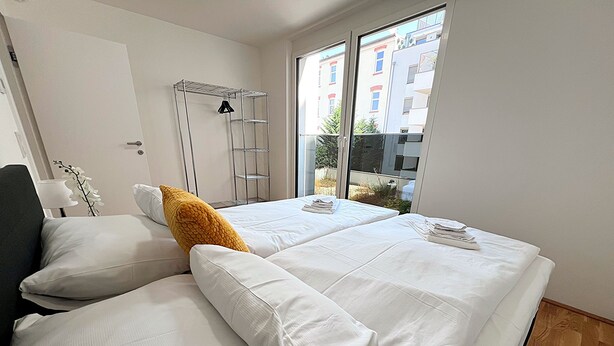 Une chambre d’hôtel avec deux lits, une fenêtre donnant sur des immeubles, un étendoir métallique et une lampe.