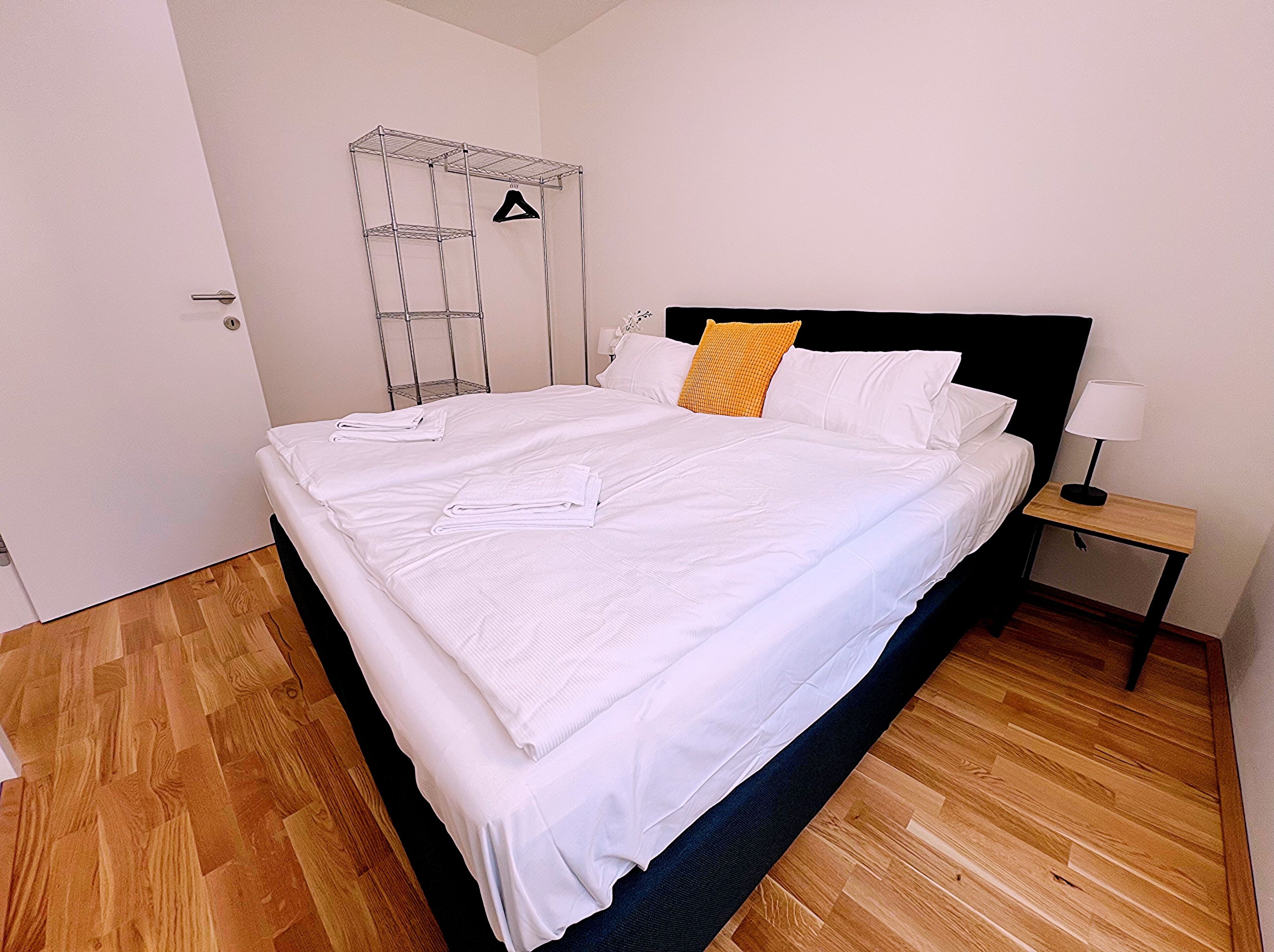 Ein ordentlich gemachtes Bett mit weißer Bettwäsche, ein Holzboden, ein Metallregal mit Kleiderhaken und ein Nachttisch mit Lampe.