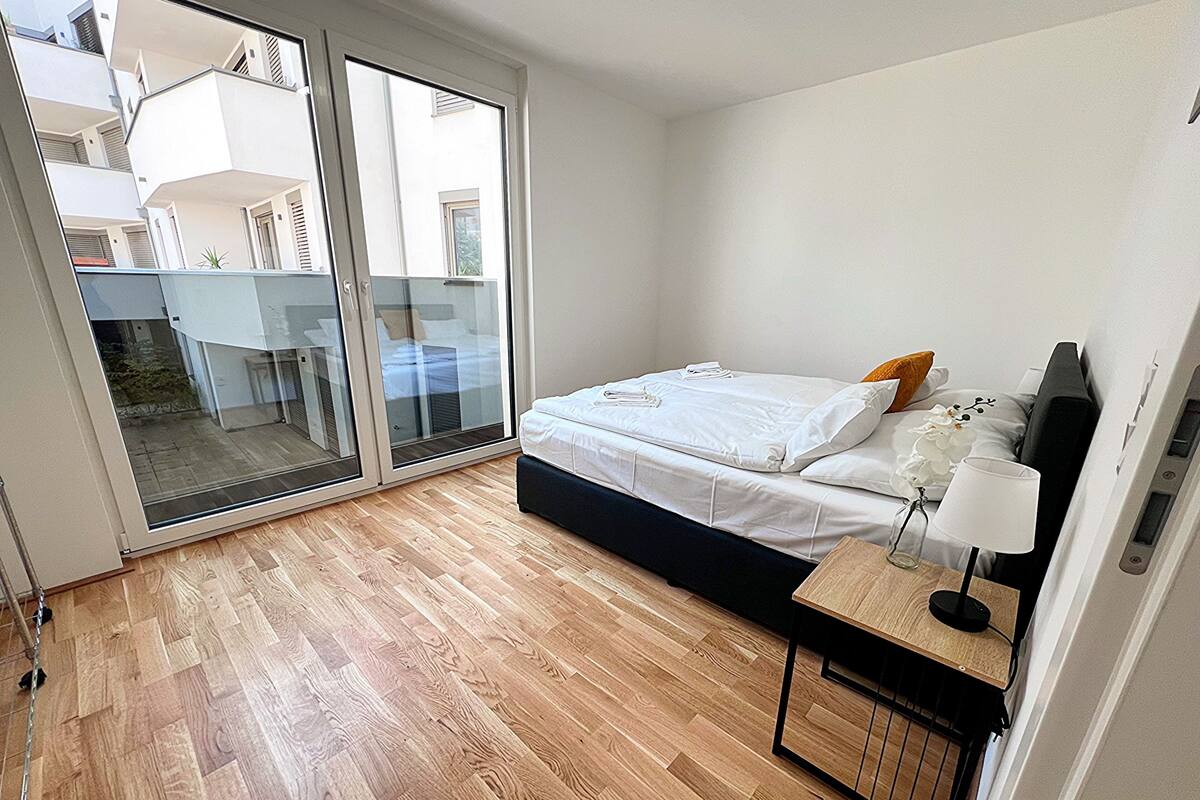 A bedroom with a bed, a bedside table with a lamp, and a sliding glass door leading to an outdoor area.