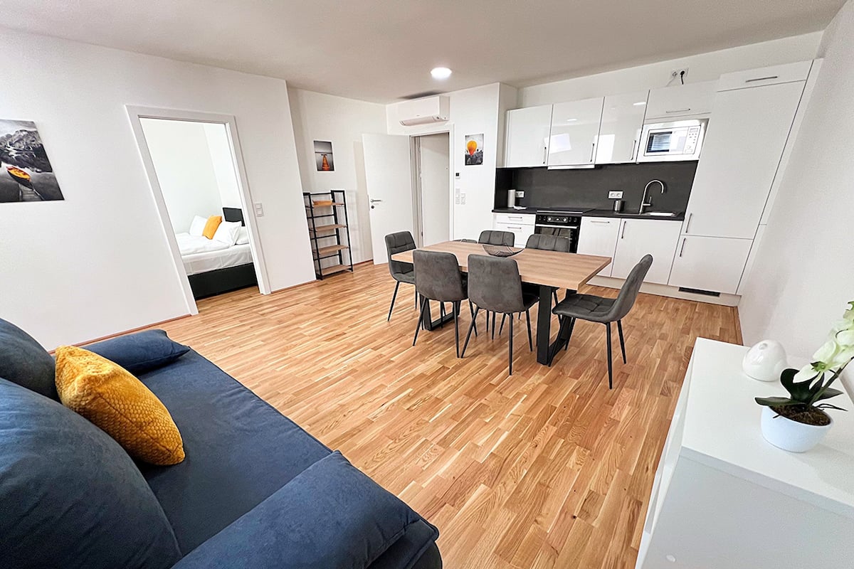 A modern apartment with a dining area, kitchenette, and a living room with a sofa and a plant.