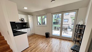 Une cuisine moderne avec des meubles blancs, un plan de travail noir et un plancher en bois. Une grande porte-fenêtre coulissante donne accès à un espace extérieur offrant une vue sur les bâtiments.