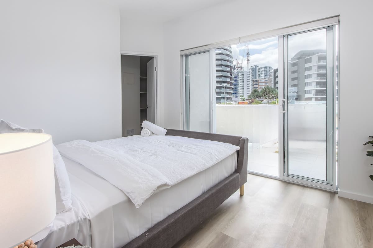 Een moderne slaapkamer met een bed, een bedlamp, een kledingkast en een glazen schuifdeur naar een balkon met uitzicht op de stad.