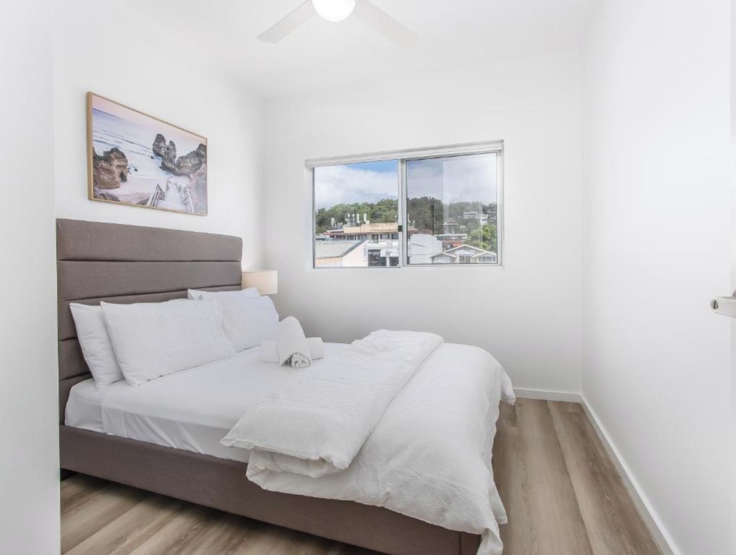 A bedroom with a bed, a window with a city view, a ceiling fan, and a framed picture on the wall.