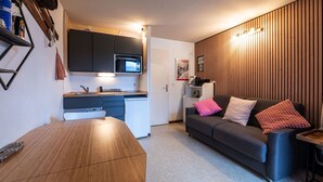 A compact living space with a kitchenette, a dining table, a sofa with cushions, and a wooden accent wall. - Lovely 4 pers studio at the foot of the pistes (Morillon)