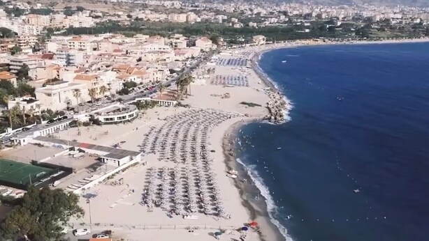 Vista aérea de una ciudad costera con playa, una fila de sombrillas y sillas de playa, un puerto deportivo y una zona urbana densamente construida al fondo.