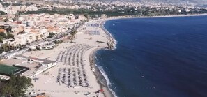 Vue aérienne d’une ville côtière, avec une plage bordée de rangées de parasols et de chaises longues, un port de plaisance, et une zone urbaine densément construite en arrière-plan.