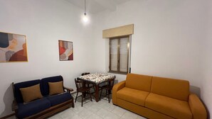 Un salón con un sofá azul, un sofá naranja, una mesa de comedor con sillas y una ventana con persianas.