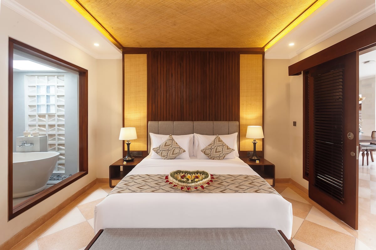 Habitación de hotel con una cama grande, dos mesitas de noche con lámparas, un espejo y vistas a un baño con bañera.