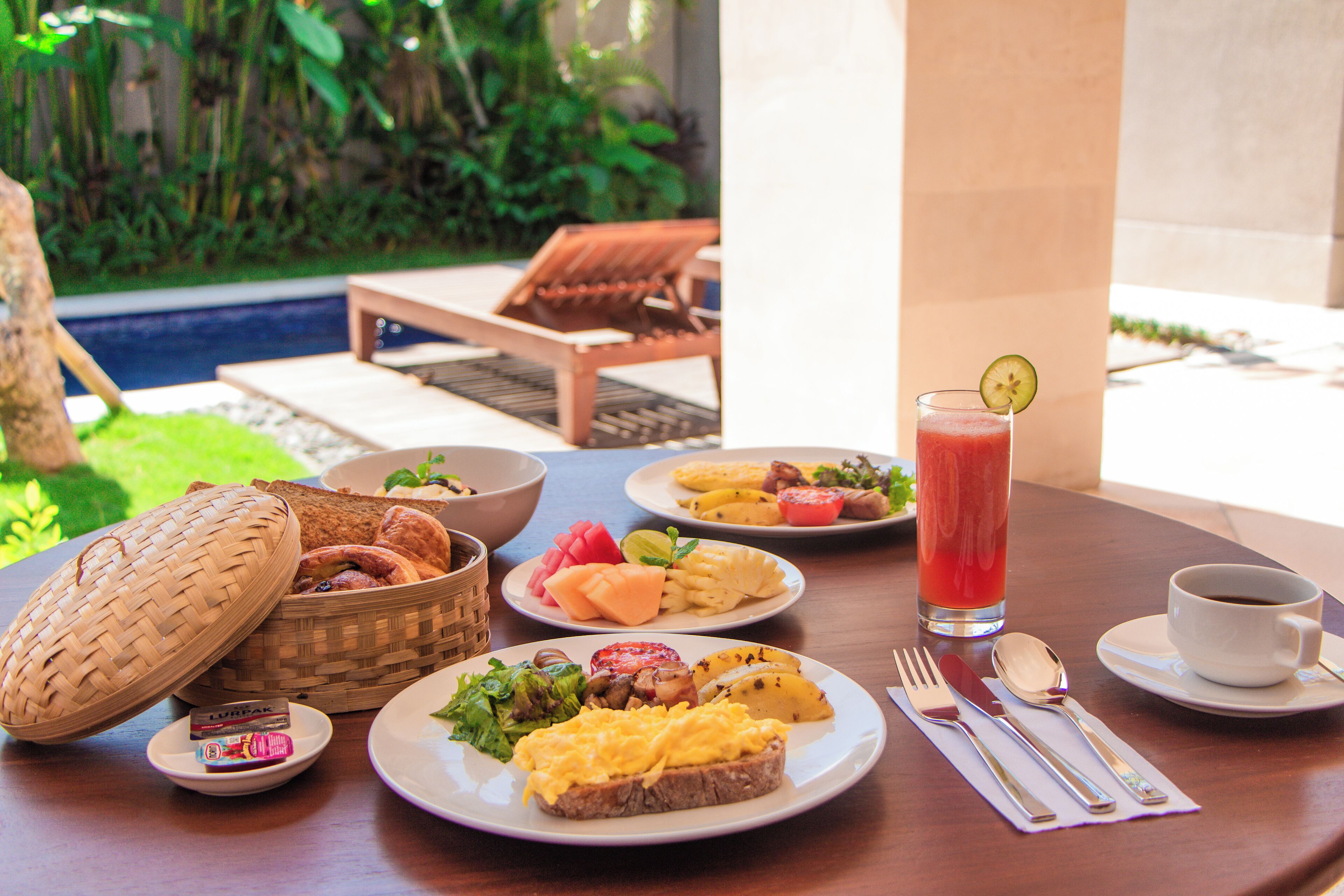 A dining table with breakfast items, a drink, and a view of a pool and greenery.