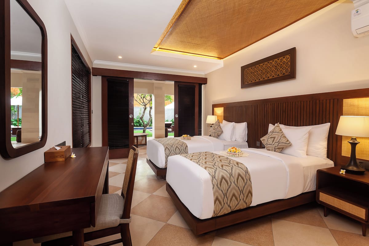 Habitación de hotel con una cama de madera, un escritorio y una silla. Hay una ventana con persianas, un aire acondicionado de pared y un adorno en la pared.