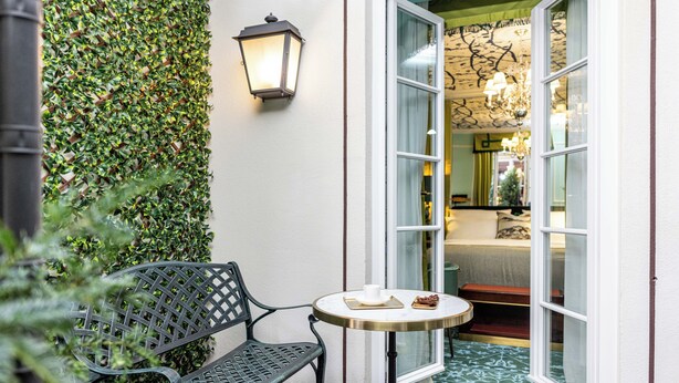 A cozy outdoor seating area with a bench and a small table, a wall-mounted lantern, and a door leading to a room with a patterned floor and a chandelier.