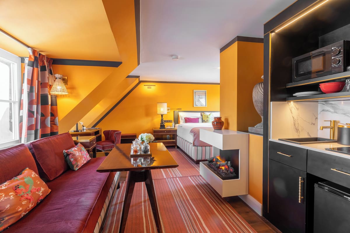 A modern hotel room with a sofa, dining table, kitchen area, and a fireplace.