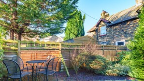 Une maison en pierre avec une clôture en bois, une terrasse avec des chaises et un jardin où poussent différentes plantes, ainsi qu'un chemin en pierre.