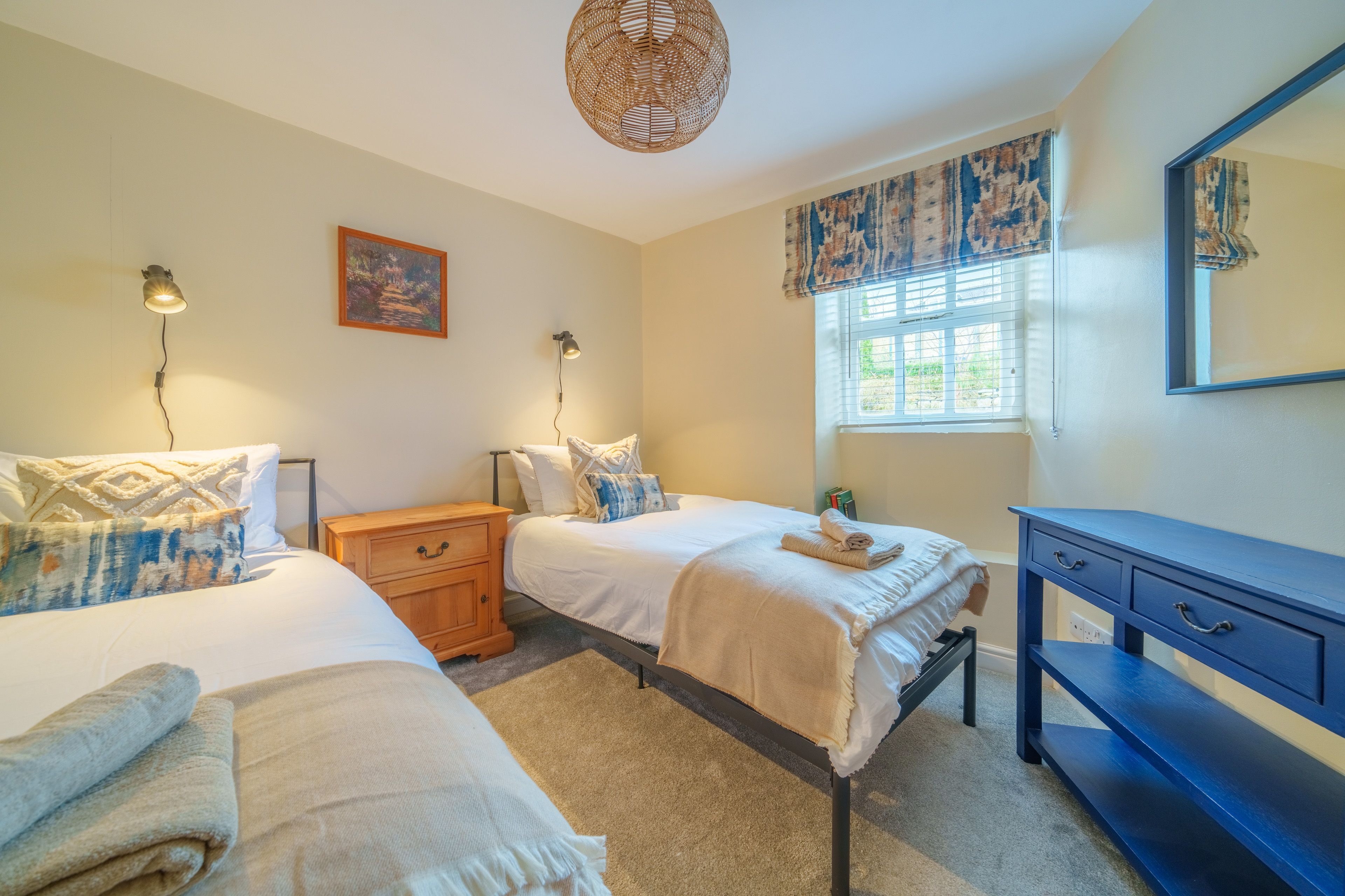 Quarto com duas camas, penteadeira, mesa de cabeceira, abajur, pintura e janela com cortinas.