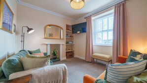 Un salon chaleureux avec un canapé vert, un fauteuil beige et un coussin rayé. On y trouve une cheminée avec un manteau de cheminée, un miroir et une étagère ornée d’objets décoratifs.