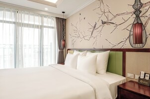 A hotel room with a large bed, a green headboard, a wall mural of branches and birds, and a nightstand with a phone and a card holder.