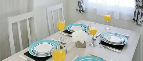 A dining table set with plates, glasses, and cutlery, with a window and curtains in the background.