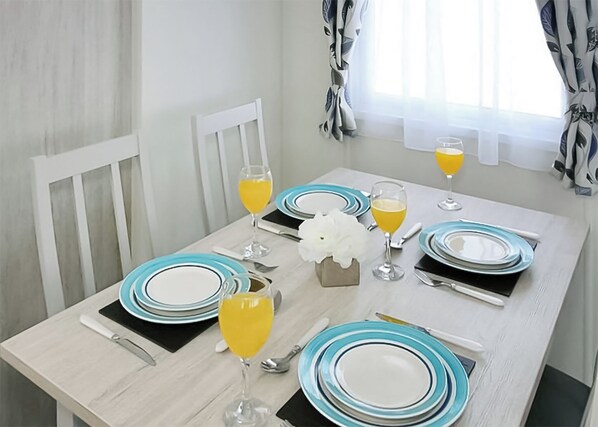 Mesa de comedor dispuesta con platos, vasos y cubiertos, con una ventana y cortinas al fondo.