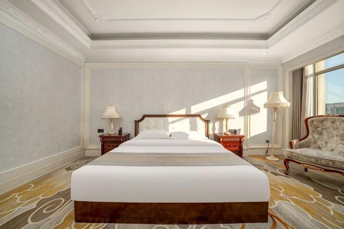 Una cama grande con cabecera de madera, dos mesitas de noche con lámparas, una silla y un ventanal con luz natural.