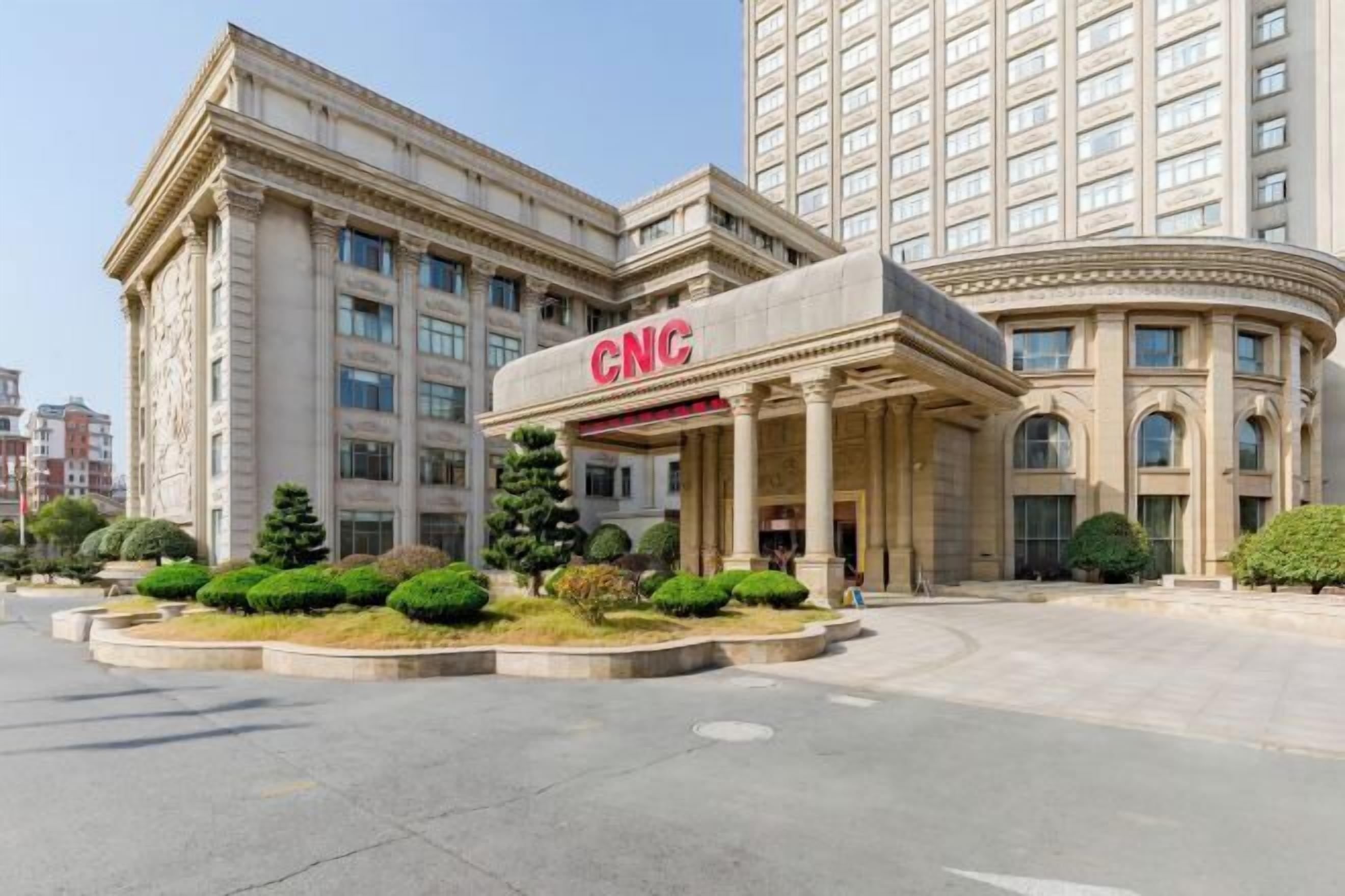 A multi-story building with a prominent CNC sign on the facade, surrounded by landscaped greenery and a clear sky.