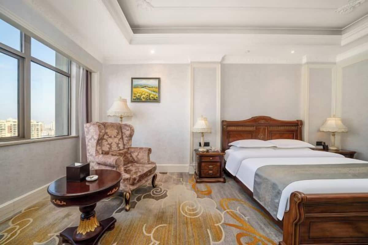 Habitación de hotel con un ventanal amplio, cama con ropa blanca, cabecera de madera, una silla, una mesita con una lámpara y un cuadro en la pared.