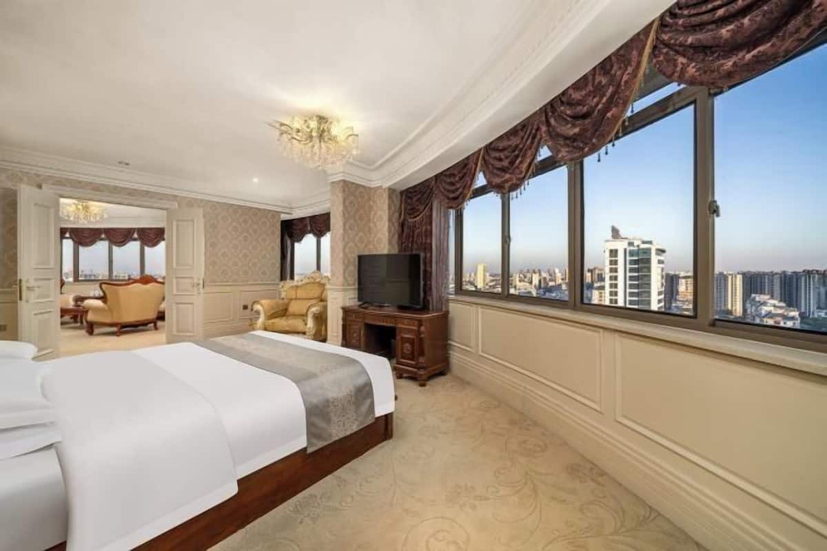 Un dormitorio amplio con una cama grande, un televisor sobre un soporte de madera y vistas de la ciudad a través de amplias ventanas.