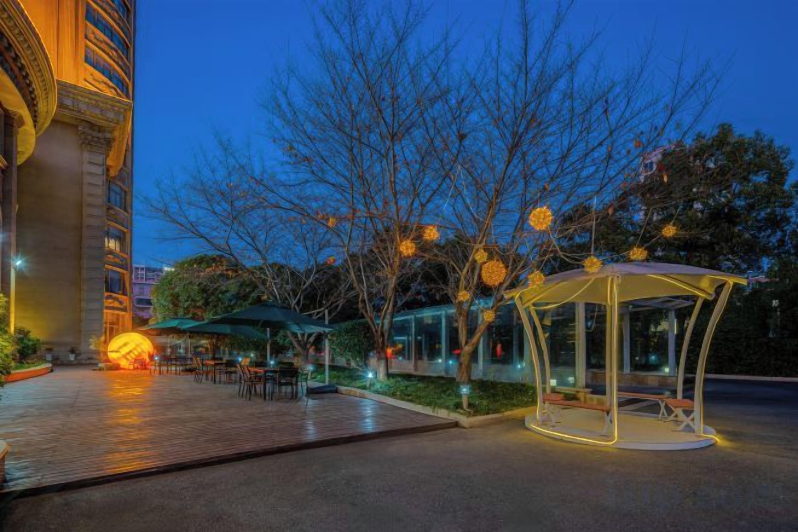 An outdoor area with a gazebo, tables, and chairs, surrounded by trees and a building.