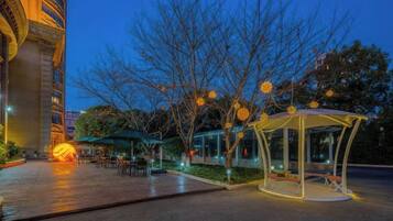 An outdoor area with a gazebo, tables, and chairs, surrounded by trees and a building.