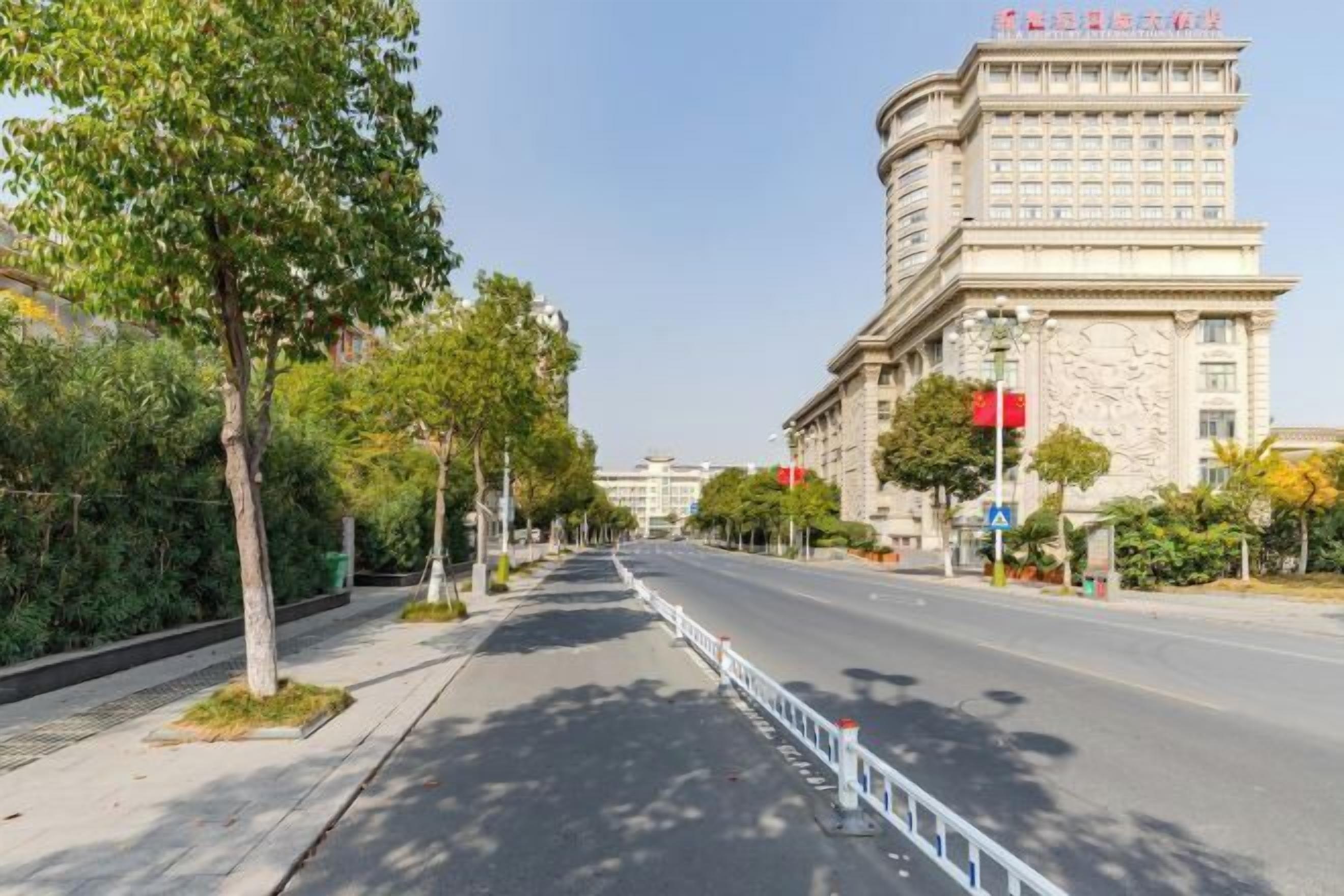 A wide, tree-lined street with a large, classical building on the right.