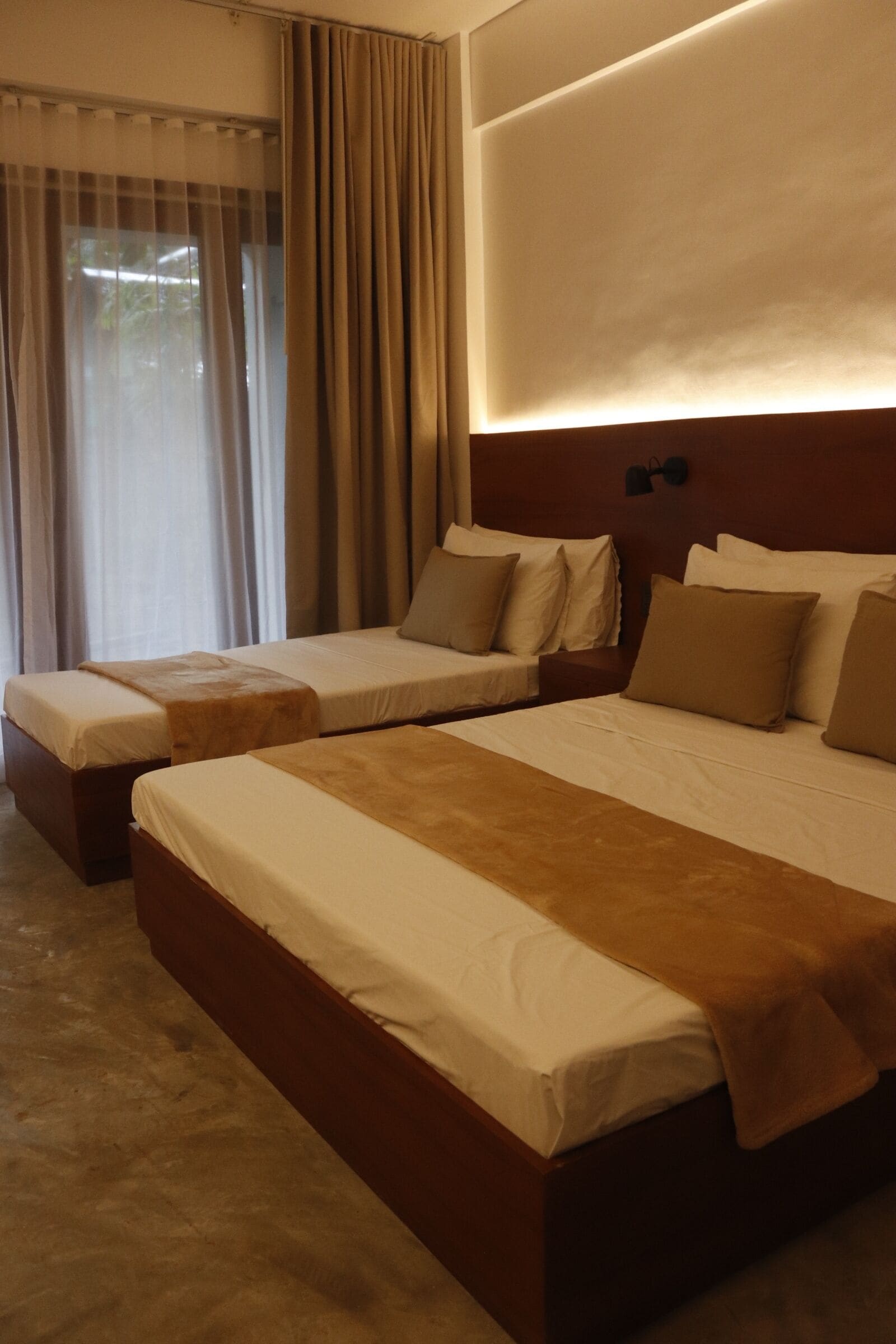 Habitación de hotel con dos camas individuales, una ventana grande con cortinas y una lámpara fijada a la pared.
