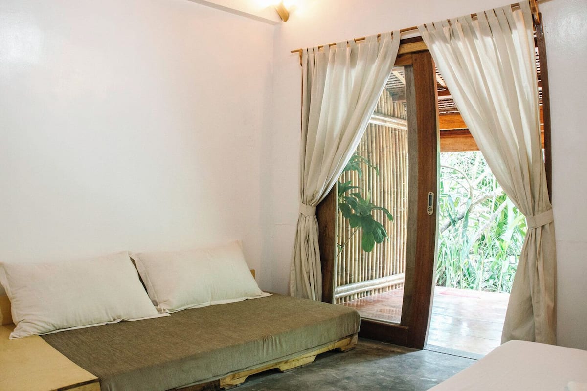 Un lit simple avec un matelas et des oreillers, une porte en bois donnant sur un paysage verdoyant et un luminaire fixé au mur.