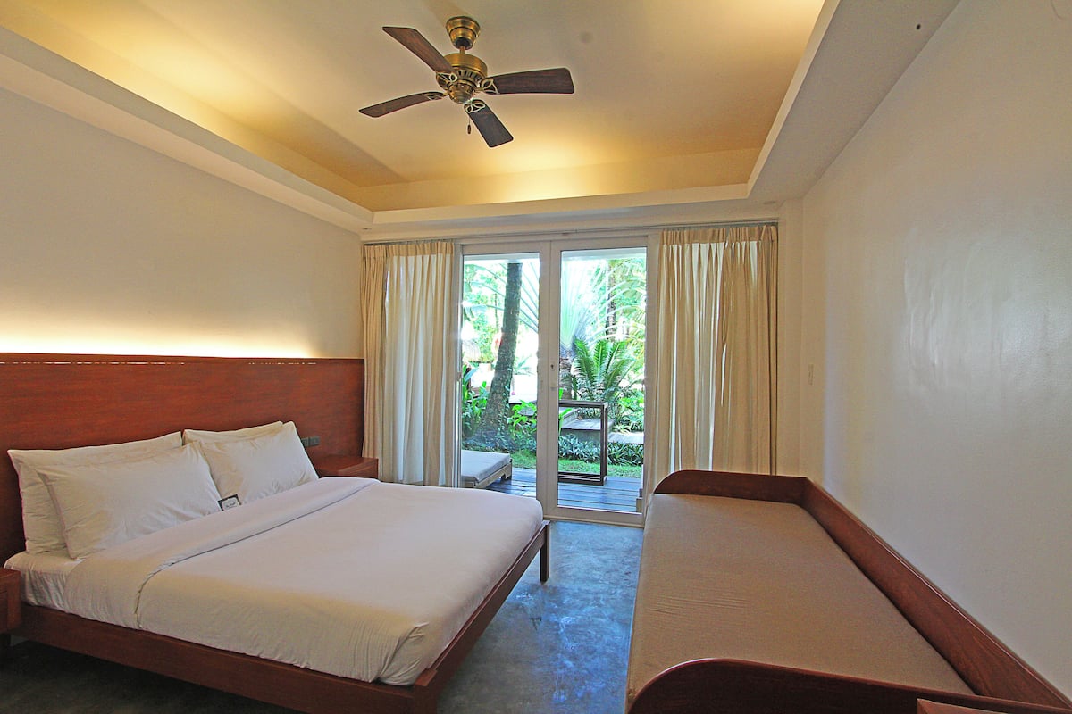 Une chambre d’hôtel avec un lit, un petit lit, un ventilateur de plafond et une vue sur la verdure par la fenêtre.