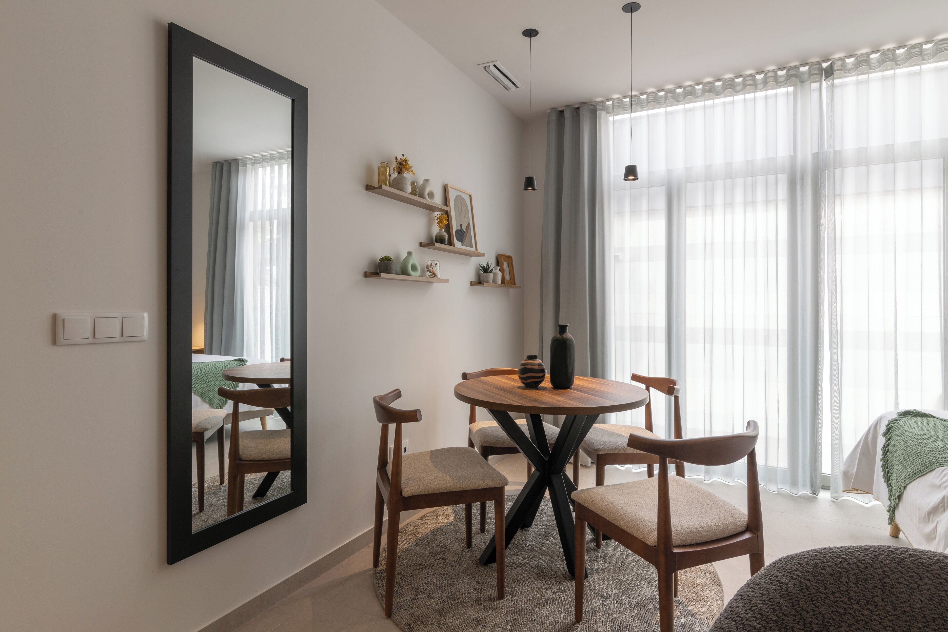 A modern living room with a round wooden table, four chairs, a large mirror, and a bed with a green blanket.