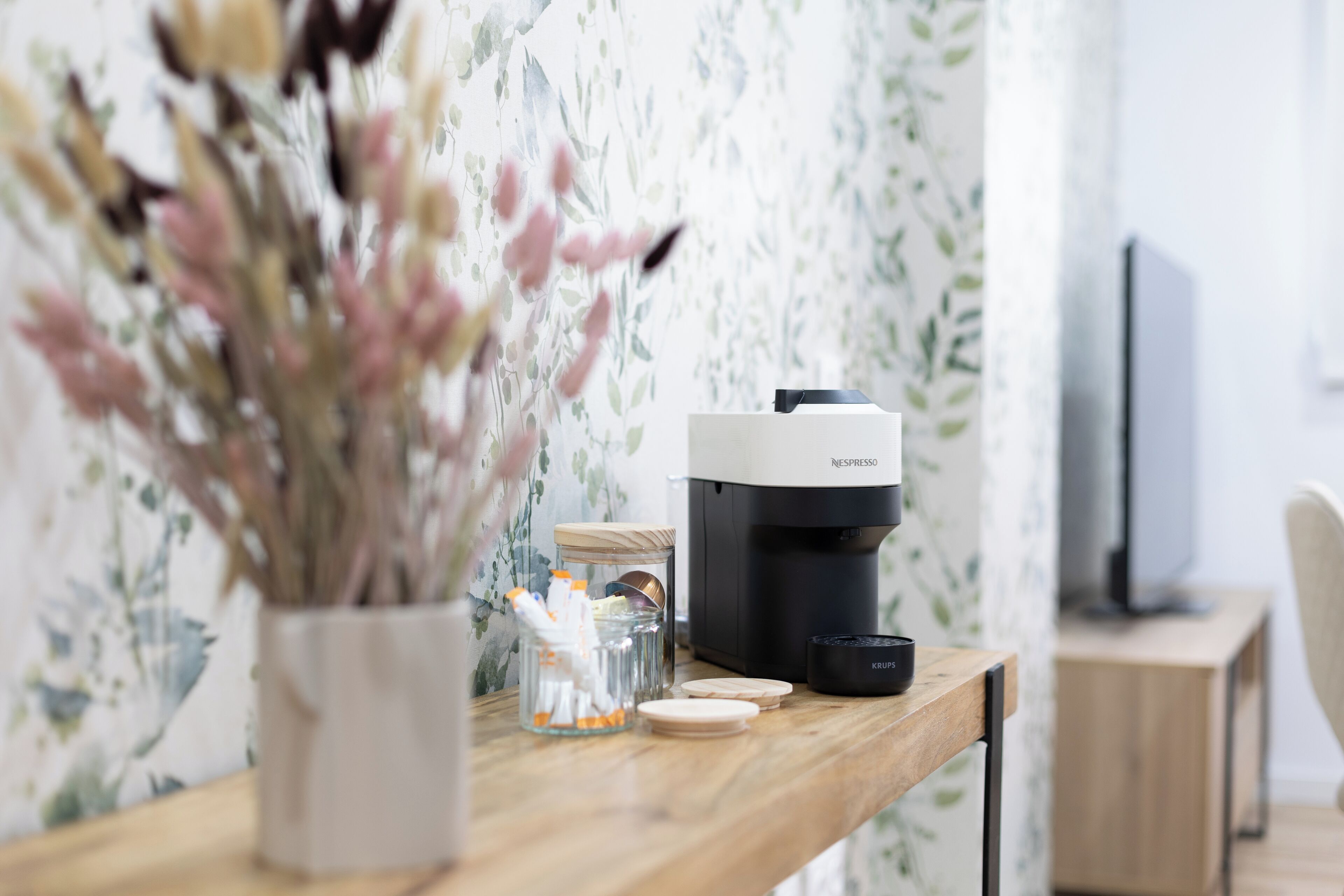 Et træbord med en Nespresso-kaffemaskine, et glas med bestik og en vase med tørrede blomster.
