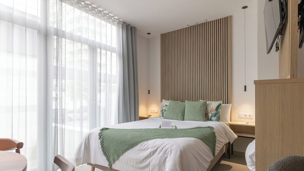 Ein modernes Hotelzimmer mit einem großen Bett, einem Holz-Kopfteil, einem Nachttisch mit Lampe und Blick ins Freie durch transparente Vorhänge.
