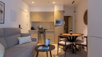 A modern living room with a grey sofa, a coffee table, and a dining area with a wooden table and chairs. A kitchen with a microwave and a dishwasher is visible in the background.