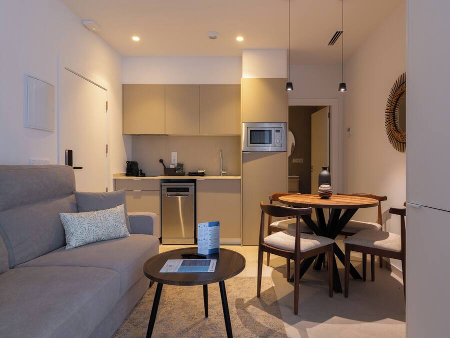 A modern living room with a grey sofa, a coffee table, and a dining area with a wooden table and chairs. A kitchen with a microwave and a dishwasher is visible in the background.