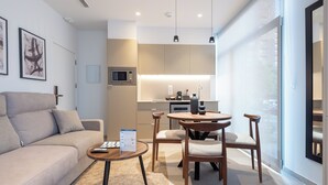 Ein modernes Wohnzimmer mit einer Couch, einem Couchtisch und einem Essbereich mit Stühlen. Es gibt eine Küchenzeile mit einer Mikrowelle und einer Kaffeemaschine. Das Zimmer hat große Fenster und bietet einen Blick ins Freie.