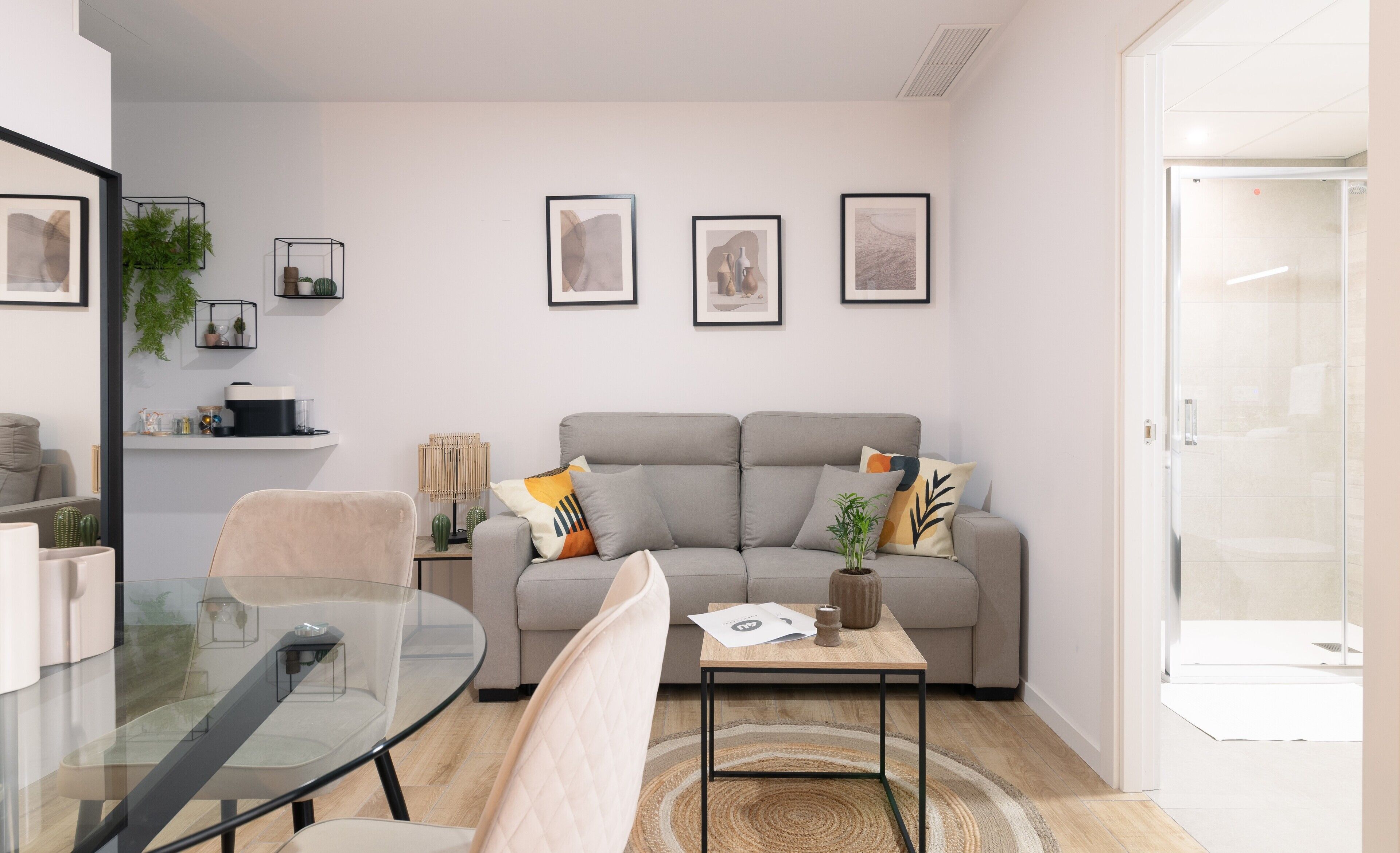 Een moderne woonkamer met een glazen tafel, een grijze bank en een bijzettafeltje met een tijdschrift en een kaars. Er hangen ingelijste foto's aan de muur en staat een plant op het nachtkastje.