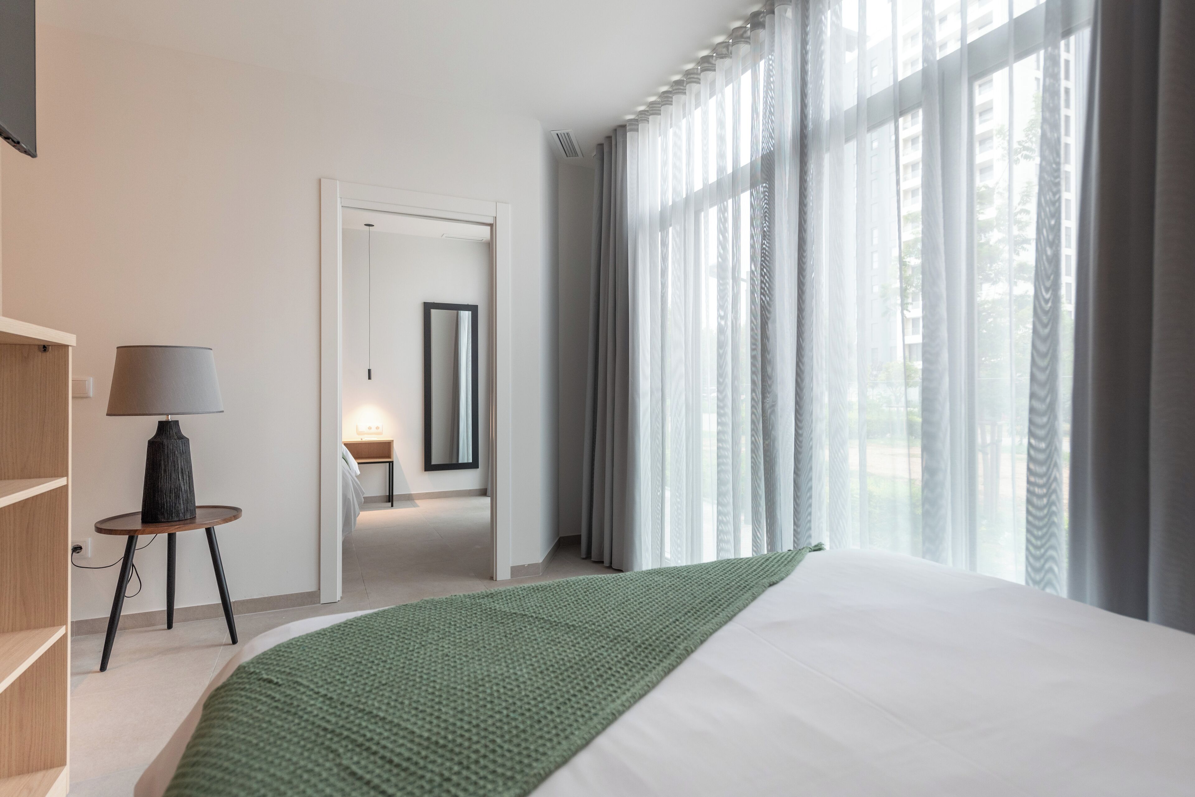 Habitación de hotel moderna con una cama grande, una mesita de noche con lámpara, vistas a través de cortinas traslúcidas y un pasillo que conduce a otra habitación.