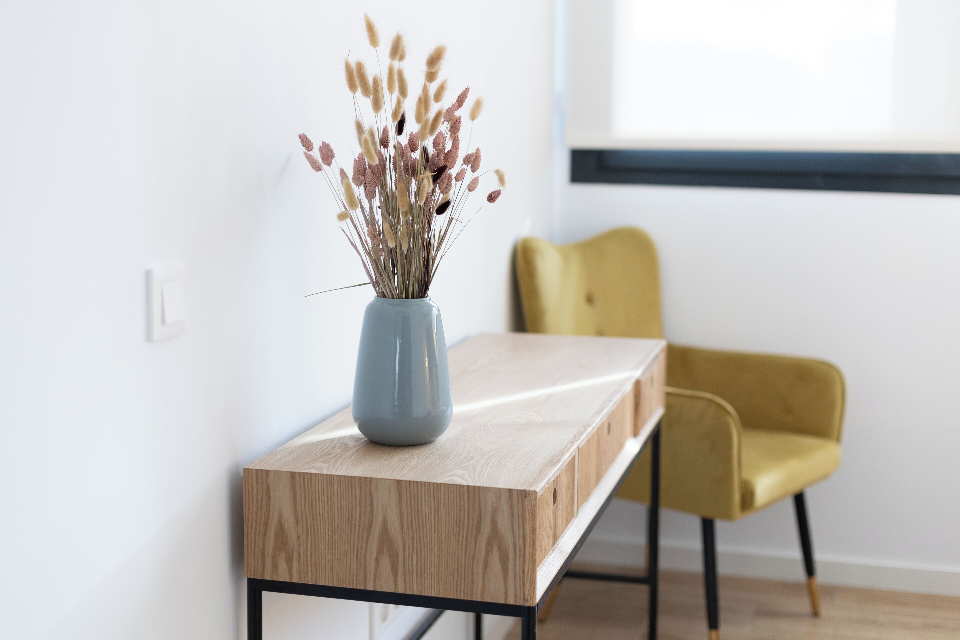 A wooden table with a vase of dried flowers, a yellow chair, and a window with blinds.
