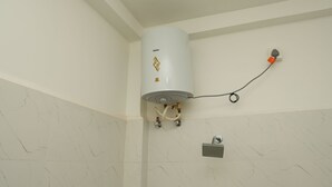 A corner bathroom with a white water heater mounted on the wall, a visible electrical outlet, and a small control panel. - Rebel De Pondicherry (Puducherry)