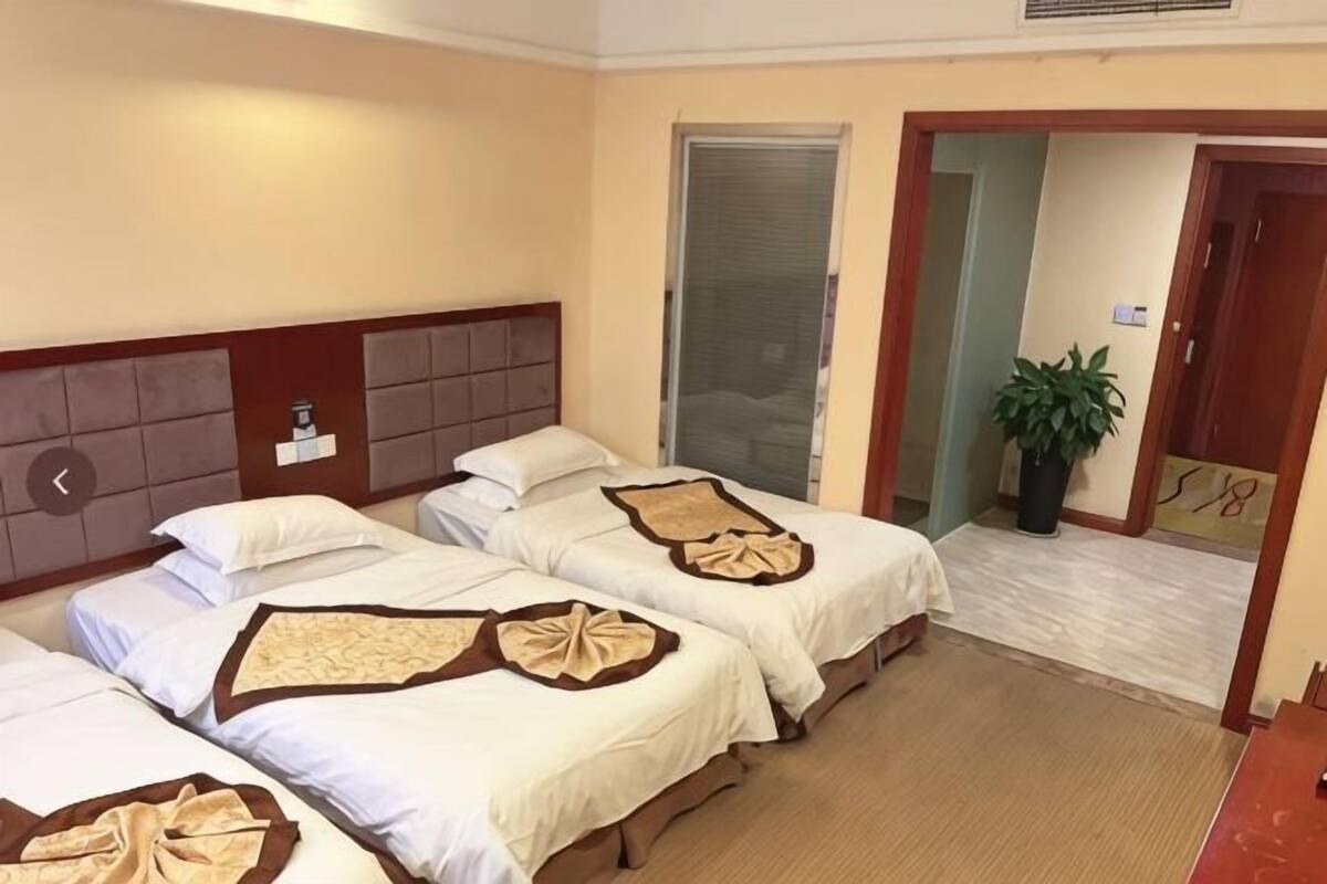 Une chambre d’hôtel avec trois lits, une tête de lit en bois, une plante en pot et une porte donnant sur une autre pièce.