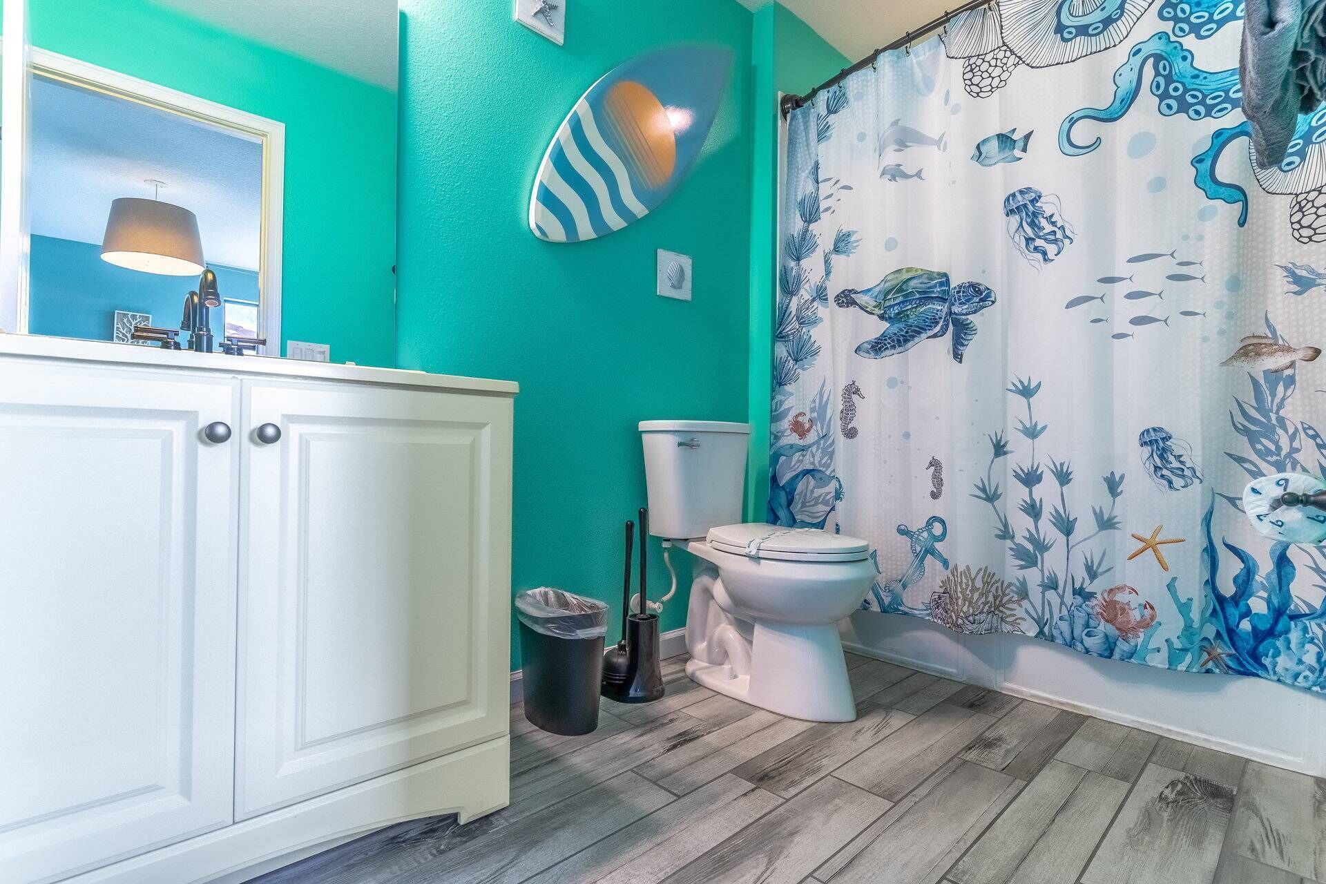 Un baño con pared verde azulado, inodoro blanco y cortina de ducha con motivos marinos.
