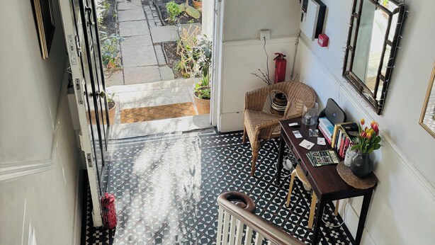 A staircase leading to a room with a patterned floor, a chandelier, a fire extinguisher, and a view of a garden through a window. - Central Victorian House (London)