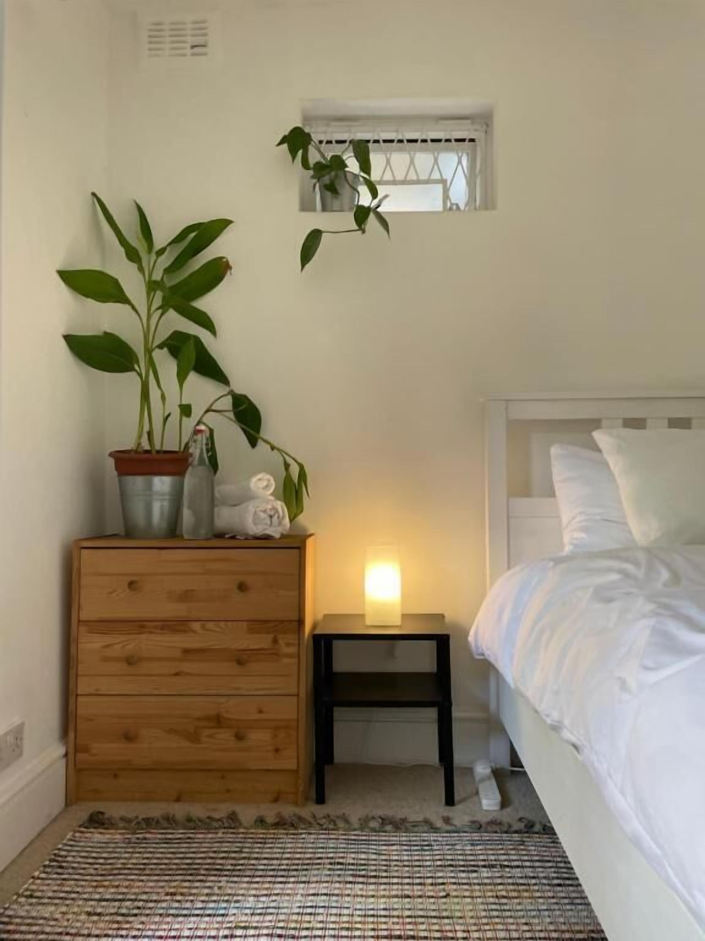 A bedroom with a bed, a bedside table with a lamp, a wooden dresser, and a potted plant.