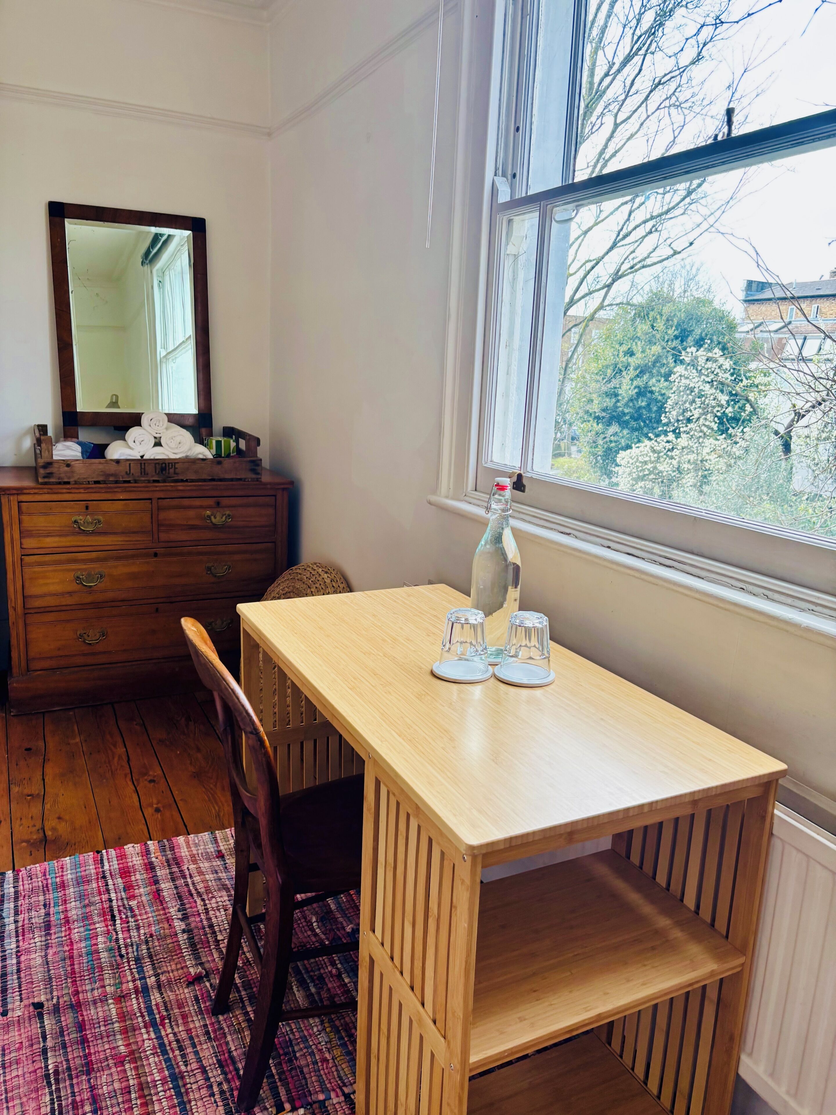 Un bureau en bois, sur lequel reposent deux verres transparents, un miroir au-dessus et une fenêtre donnant sur des arbres.